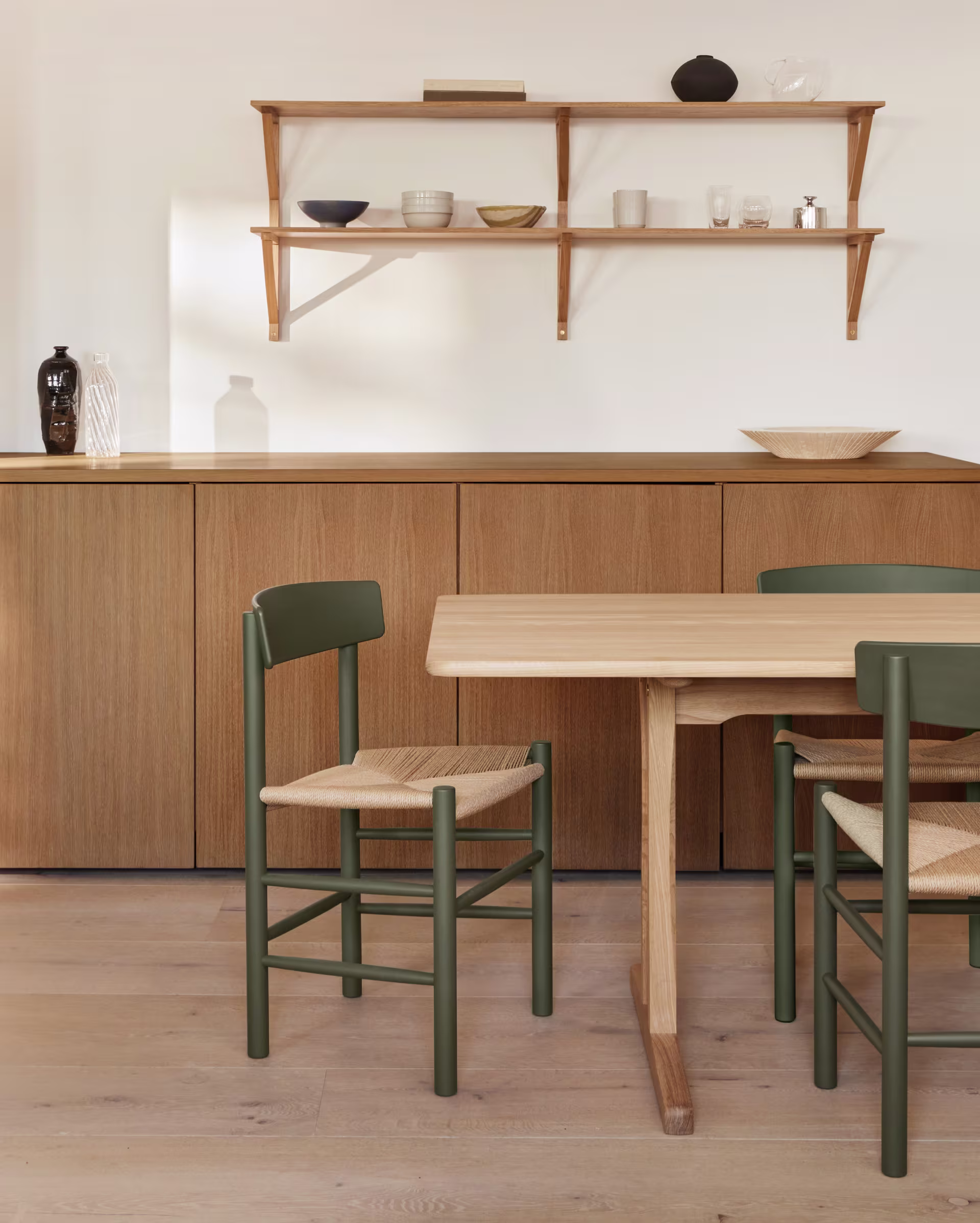 Et trebord med J39-stoler fra Fredericia Furniture med mørkegrønne rammer og flettede seter. En lav trekkommode er synlig i bakgrunnen.