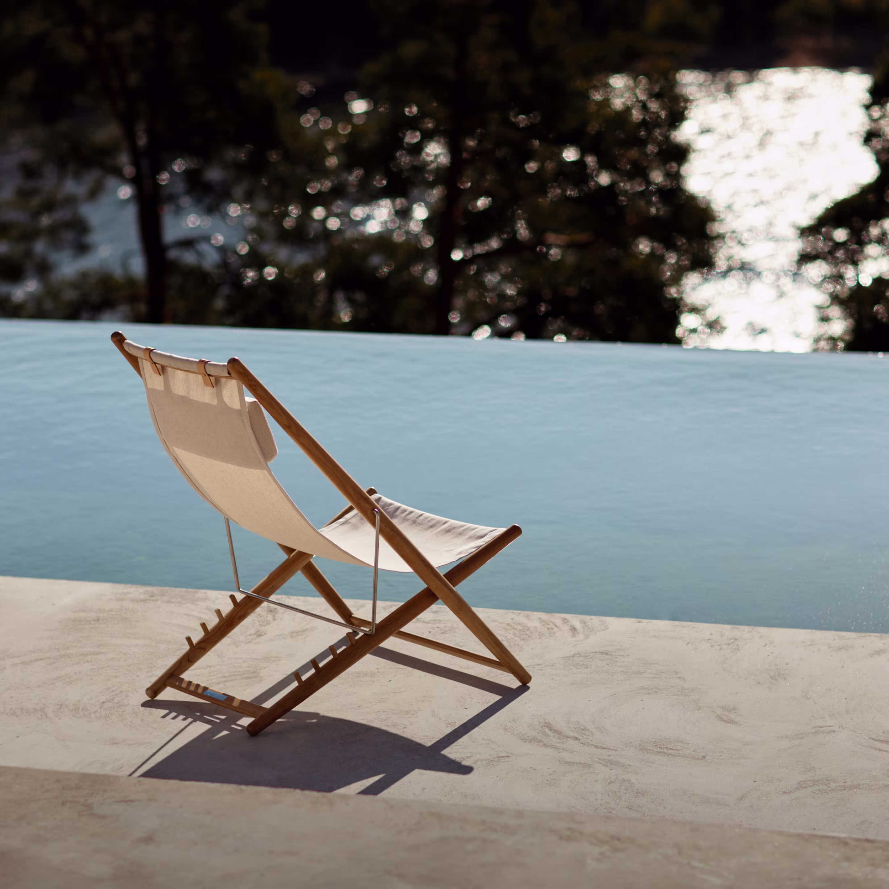En solstol med träram och beige tyg står vid kanten av en infinitypool. Vattnet i poolen är ljust blått och reflekterar ljus. I bakgrunden syns träd och en glittrande vattenyta.
