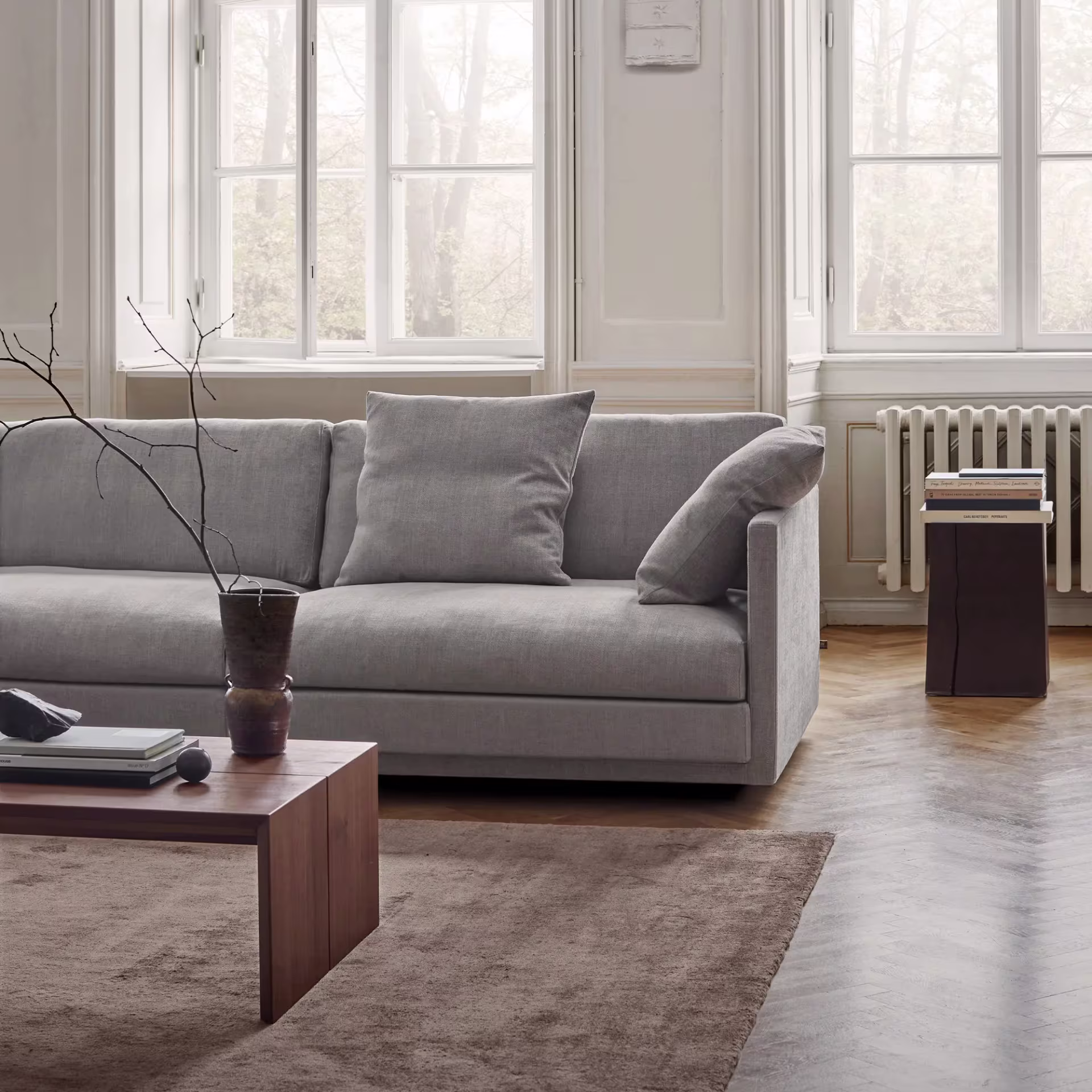 Et lyst og romslig stuemiljø med en stor, lysegrå Eilersen sofa. Foran sofaen står et rektangulært salongbord i mørkt tre med en vase med tørkede grener og noen interiørdetaljer. Gulvet er et fiskebensparkettgulv dekket av et mykt, brunt teppe. Store vinduer med hvite karmer slipper inn naturlig lys.