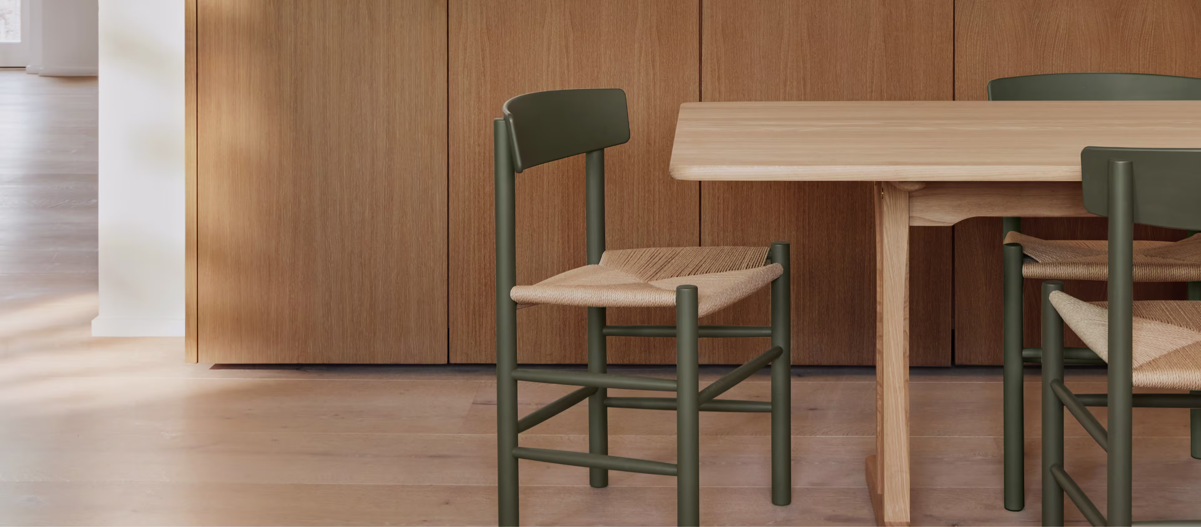 Et trebord med J39-stoler fra Fredericia Furniture med mørkegrønne rammer og flettede seter. En lav trekkommode er synlig i bakgrunnen.