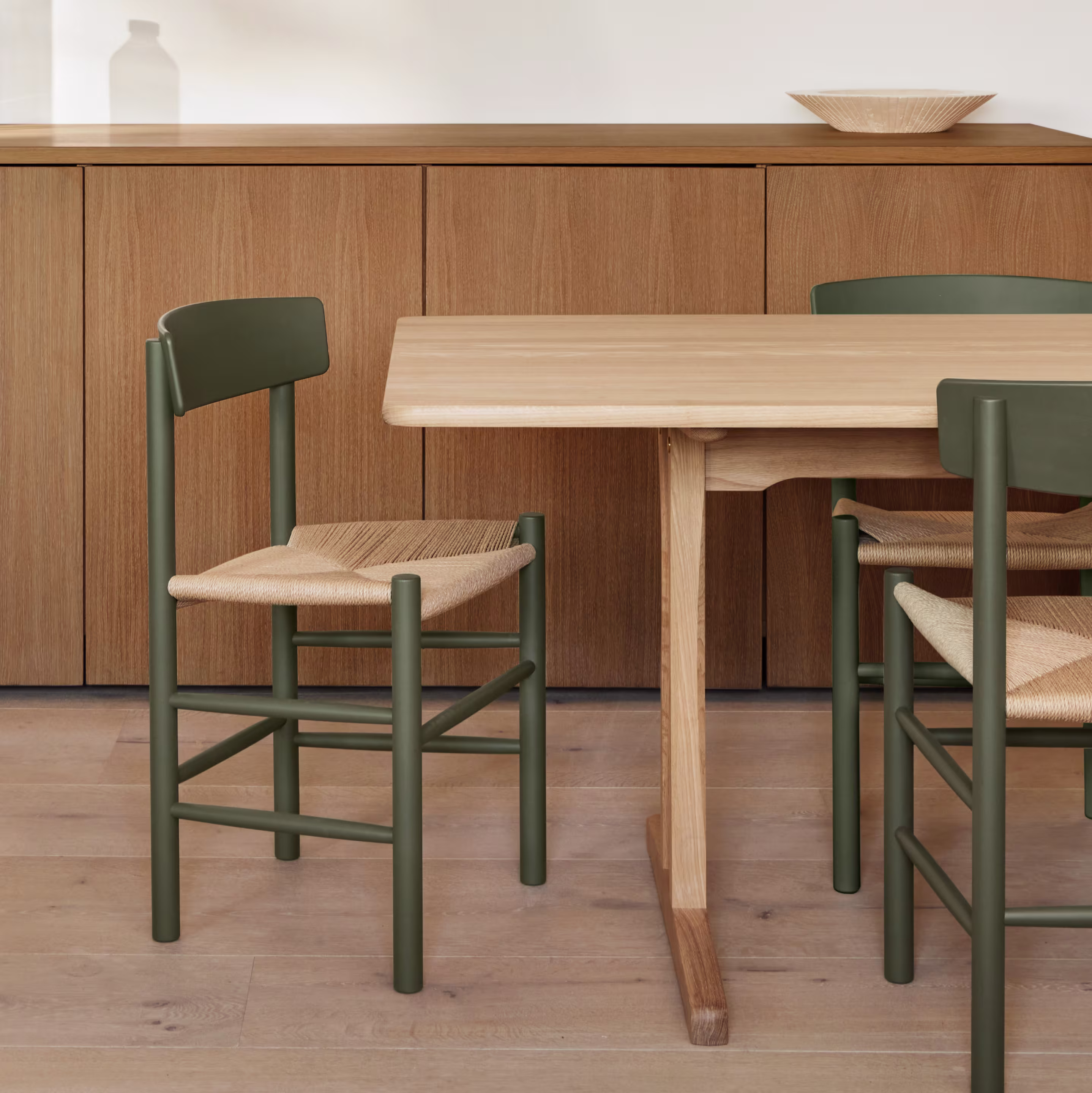 Ett träbord med J39-stolar från Fredericia Furniture med mörkgröna ramar och flätade sitsar. En låg träbyrå syns i bakgrunden.