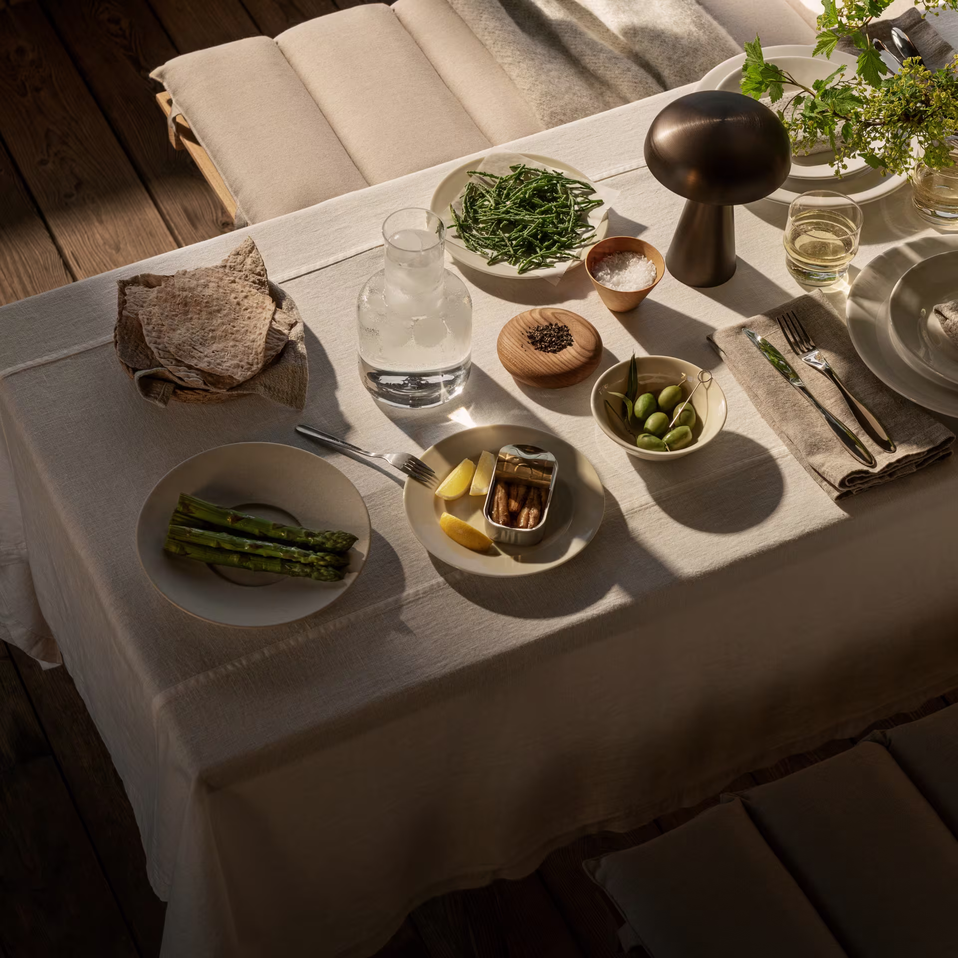 Sommerdekket bord med asparges, kokte poteter, brød, sardiner, grønne oliven og en karaffel med isvann i varm kveldssol.