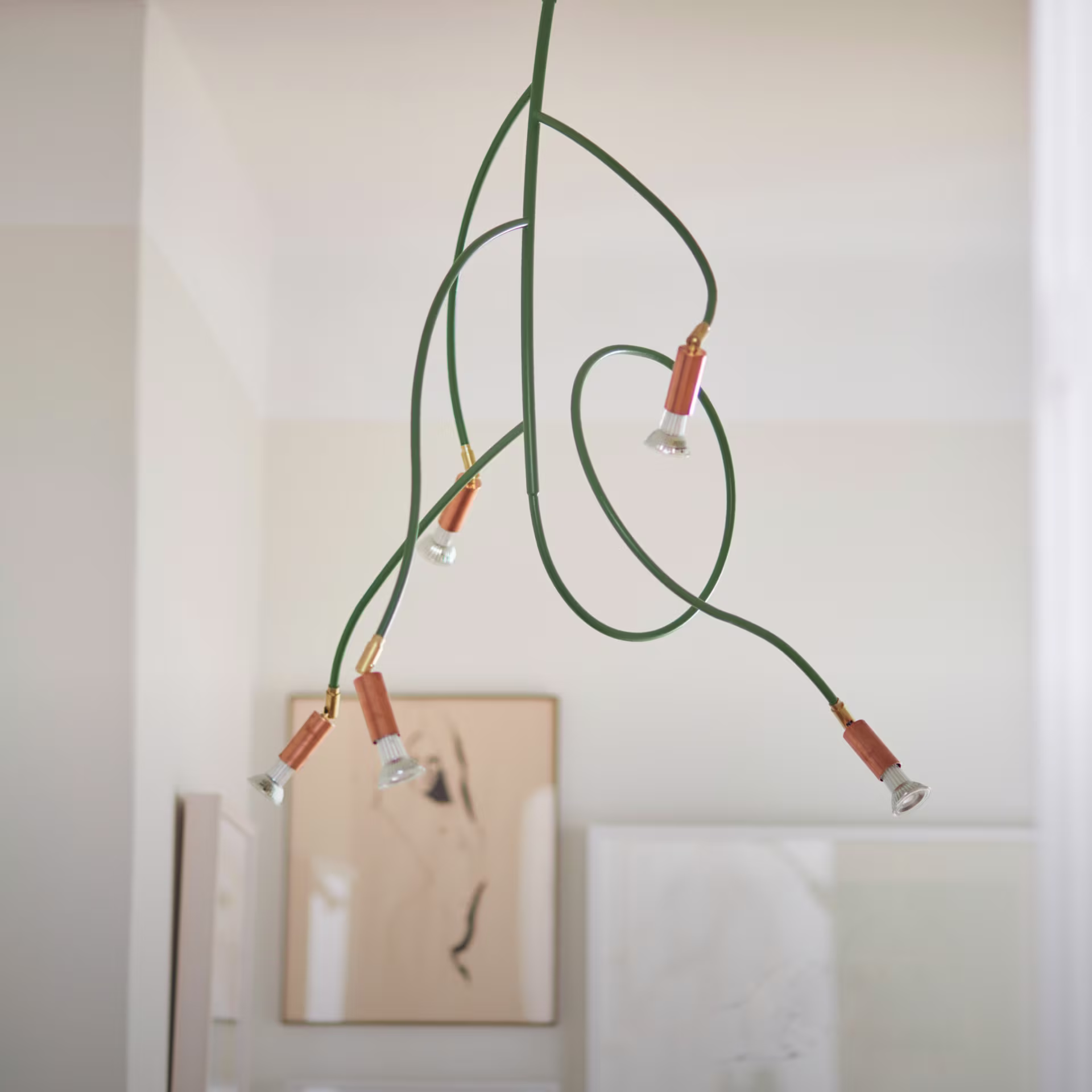 Örsjö Belysning Kvist taklampa med gröna, böjda armar och flera ljuskällor med kopparfärgade detaljer, hängande mot en ljus vägg. I bakgrunden syns delar av en tavelram.
