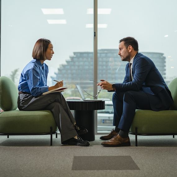 Two diverse professionals in a modern office engaged in a serious interview or meeting, one speaking intently while the other listens and takes notes, emphasizing communication and teamwork