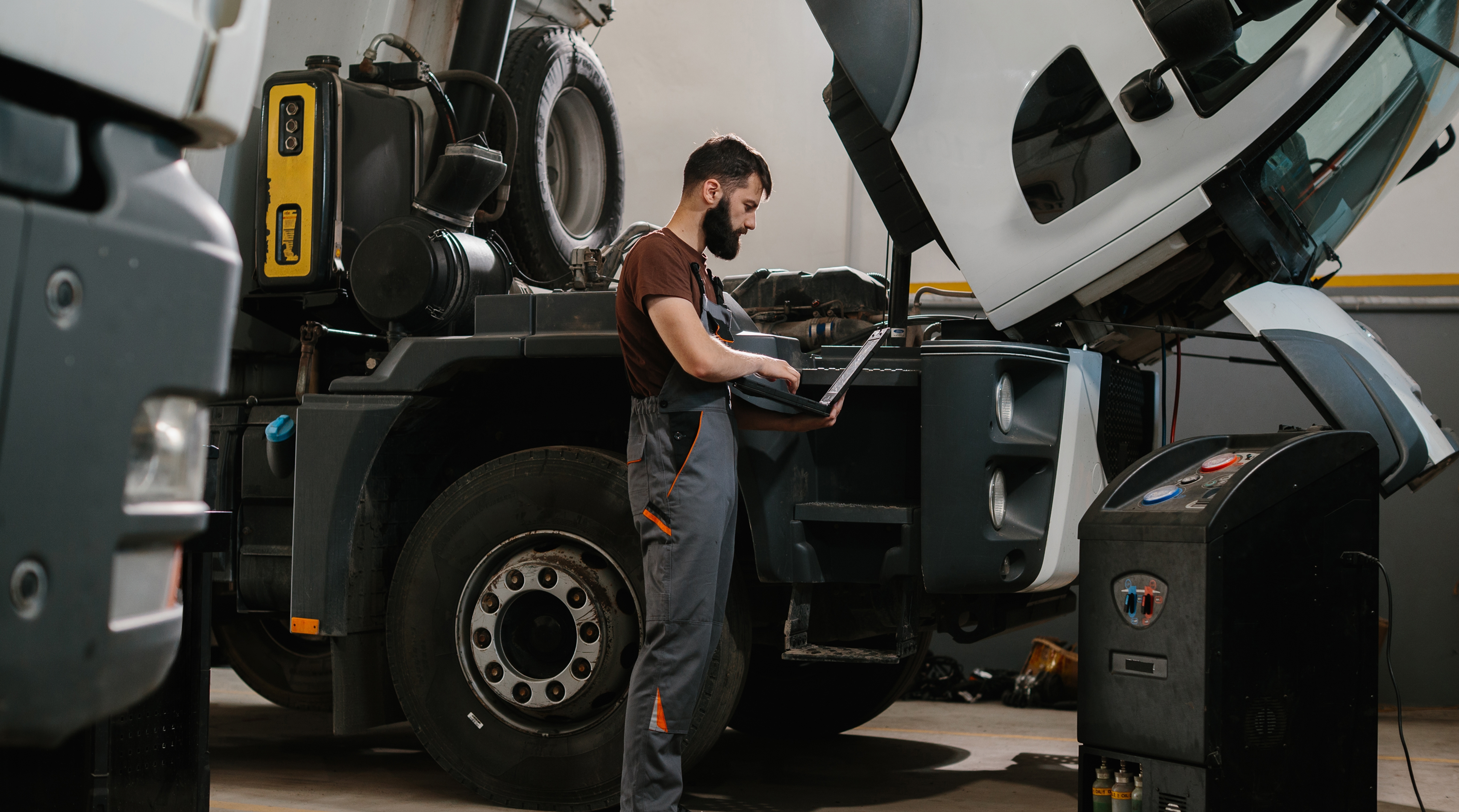 Auto mechanic working on diagnostics with a laptop while repairing a semi-truck inside a garage, focused on vehicle maintenance tasks
