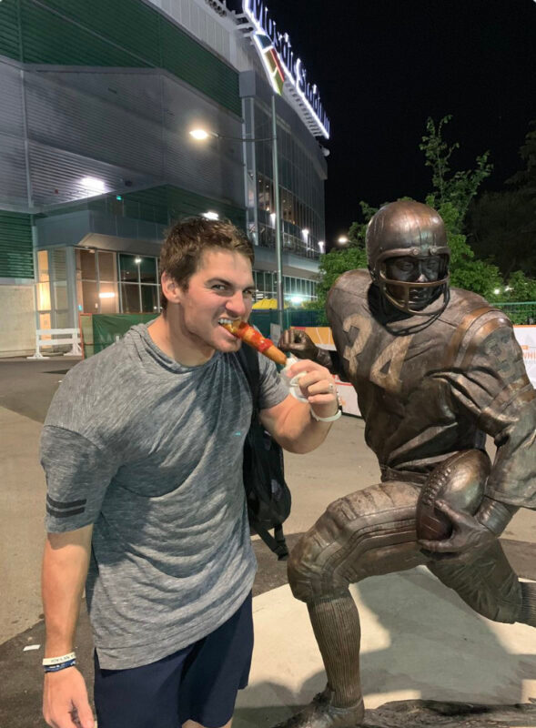 Cody Fajardo eating a corn dog while standing next to the George Reed statue at Mosaic Stadium