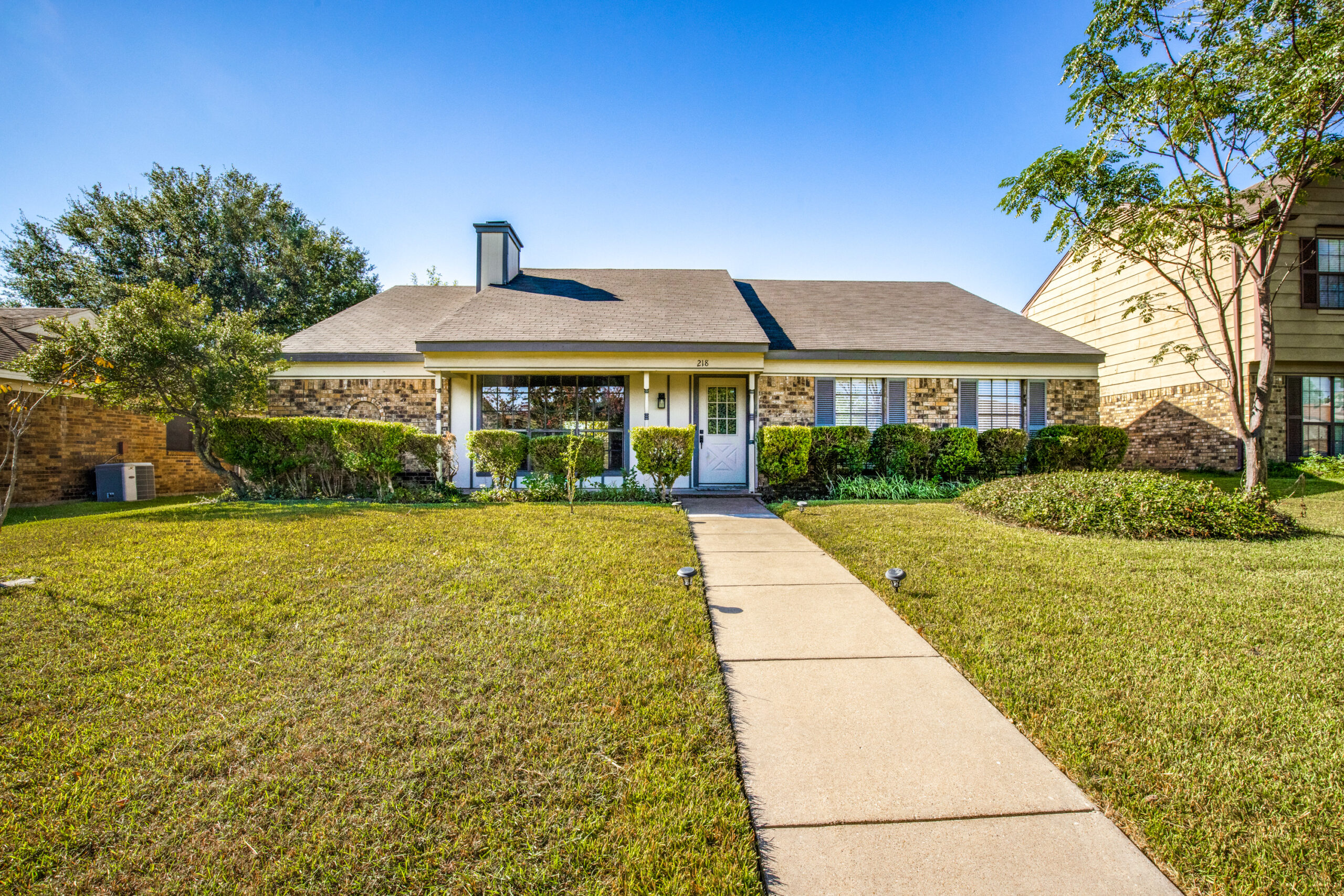 Older ranch-style home with multi-colored brown brick, a brown roof, blue shutters, dead grass, and a sidewalk leading up to the front door.