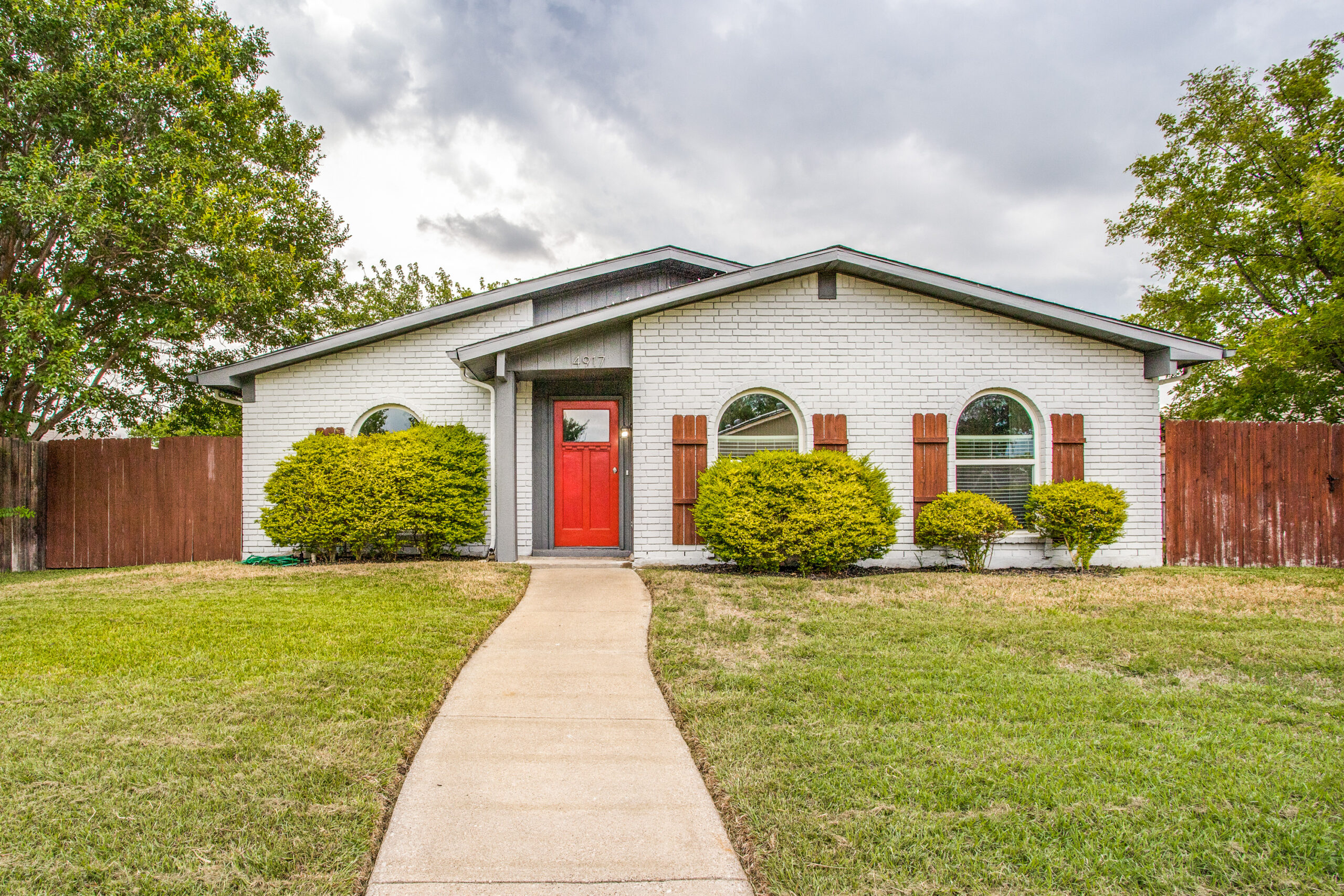 Photo of residential one-store home with white brick, wood shutters, a bright-red door, sidewalk to the front door with bushes on either side, and green/brown lawn.