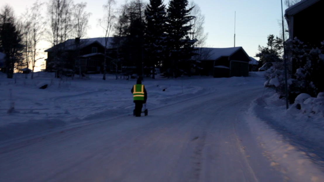 Person i reflexväst drar en barnvagn på en snöig väg i mörker.