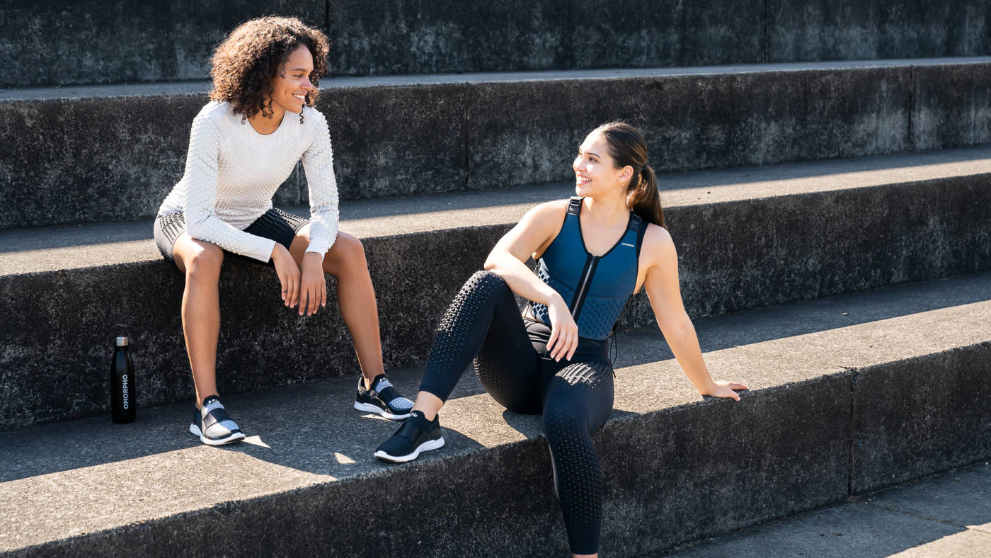Two women sitting on steps wearing OMORPHO weighted workout vest and weighted biker shorts for women on a sunny day, post workout. 