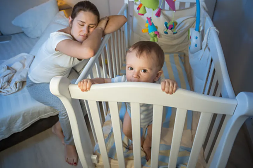Tired mother asleep next to awake baby's crib.