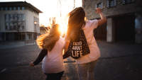 两名年轻女子跳在一起，一个穿着背包，上面写着兴奋LIFE