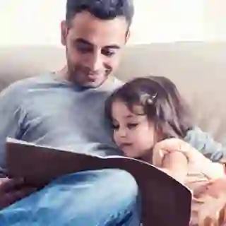 Father reading book to young daughter on couch.