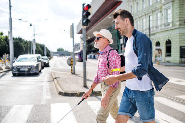 年轻人和盲目的高级白色手杖行走在城市，过马路
