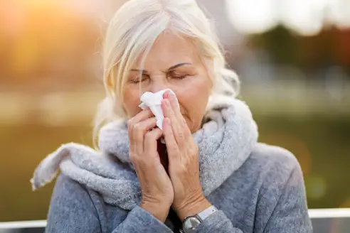 Close-up of a mature woman wearing a scarf outdoors, blowing her nose into a tissue.