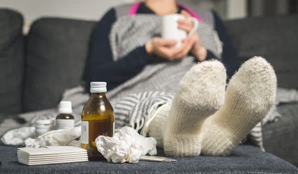 一个生病的女人躺在沙发上，喝着茶，吃着感冒药，还有一张揉皱的面巾纸。