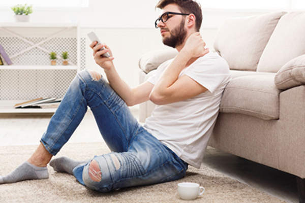 人在家里坐在地板上,手机上,揉他的脖子。