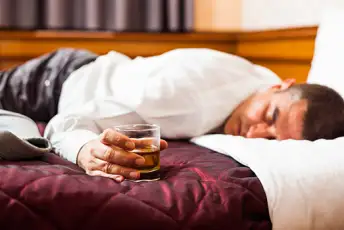 一名男子面朝下躺在床上，手里拿着一杯威士忌。
