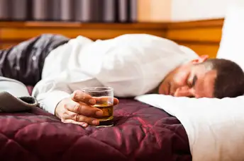 一个男人脸朝下趴在床上睡着了，手里拿着一杯威士忌。
