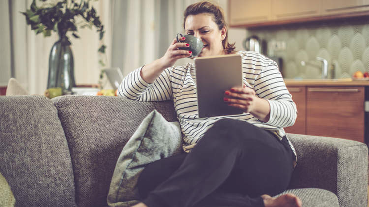超重妇女看平板电脑在家的沙发上喝茶。