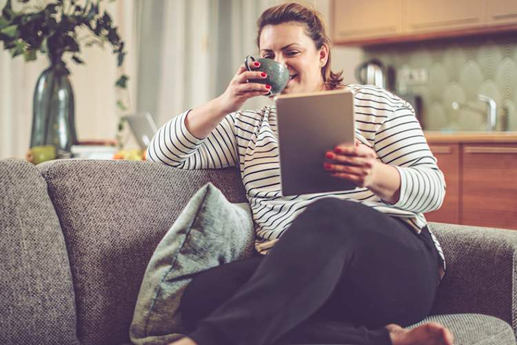 超重妇女看平板电脑在家的沙发上喝茶。