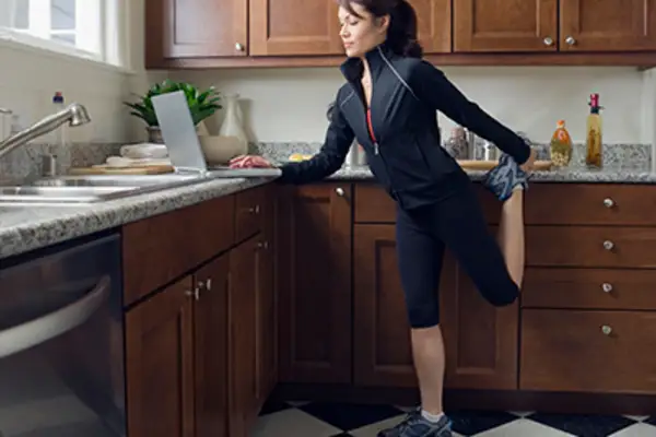 一位女士在厨房里一边看笔记本电脑一边伸懒腰。