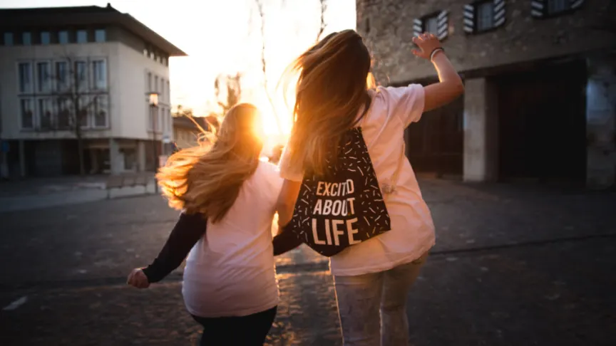 两个年轻女子跳在一起，一个背着写着“对生活感到兴奋”的背包