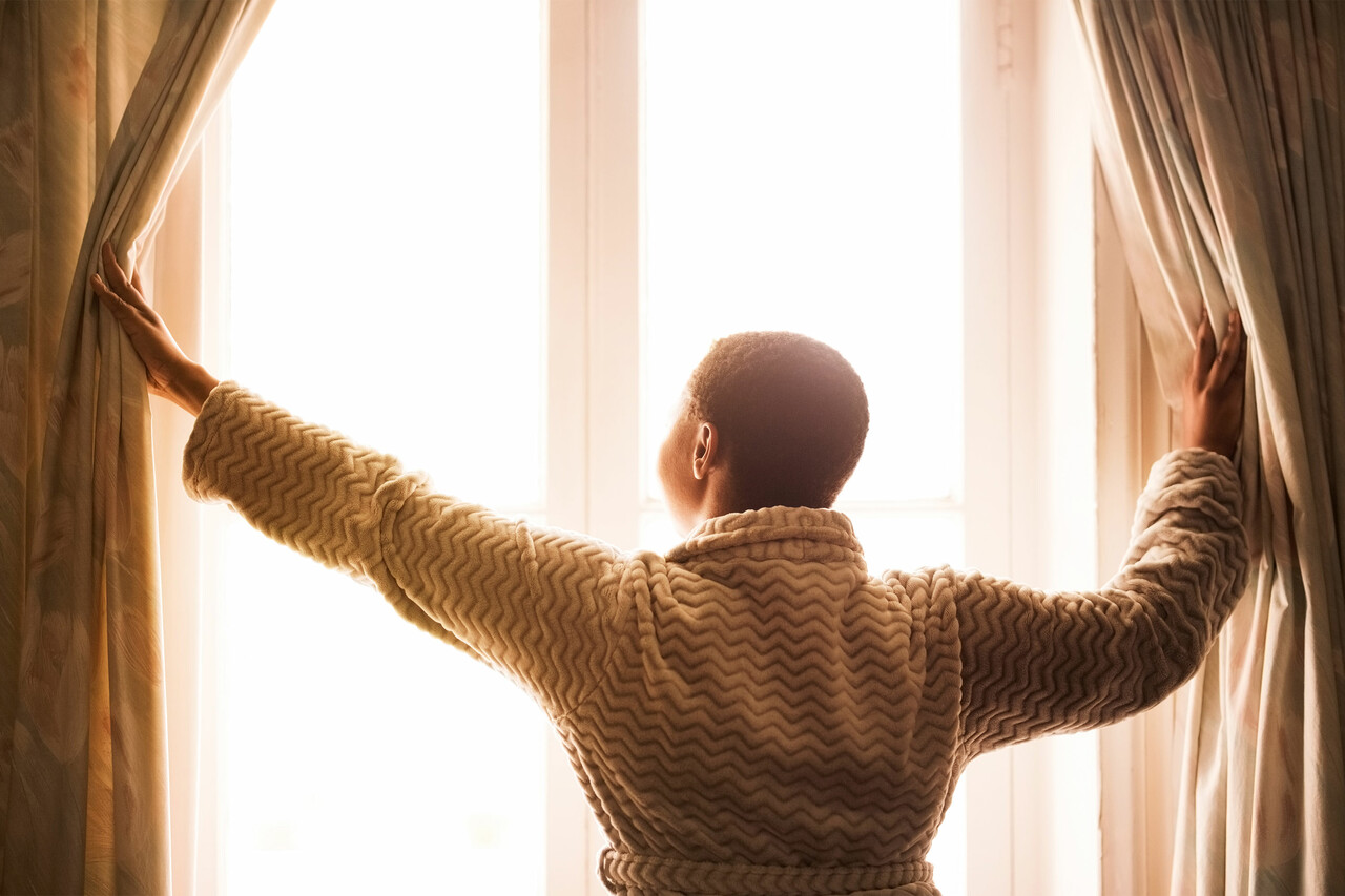 A woman in a bathrobe opens her curtains in the morning