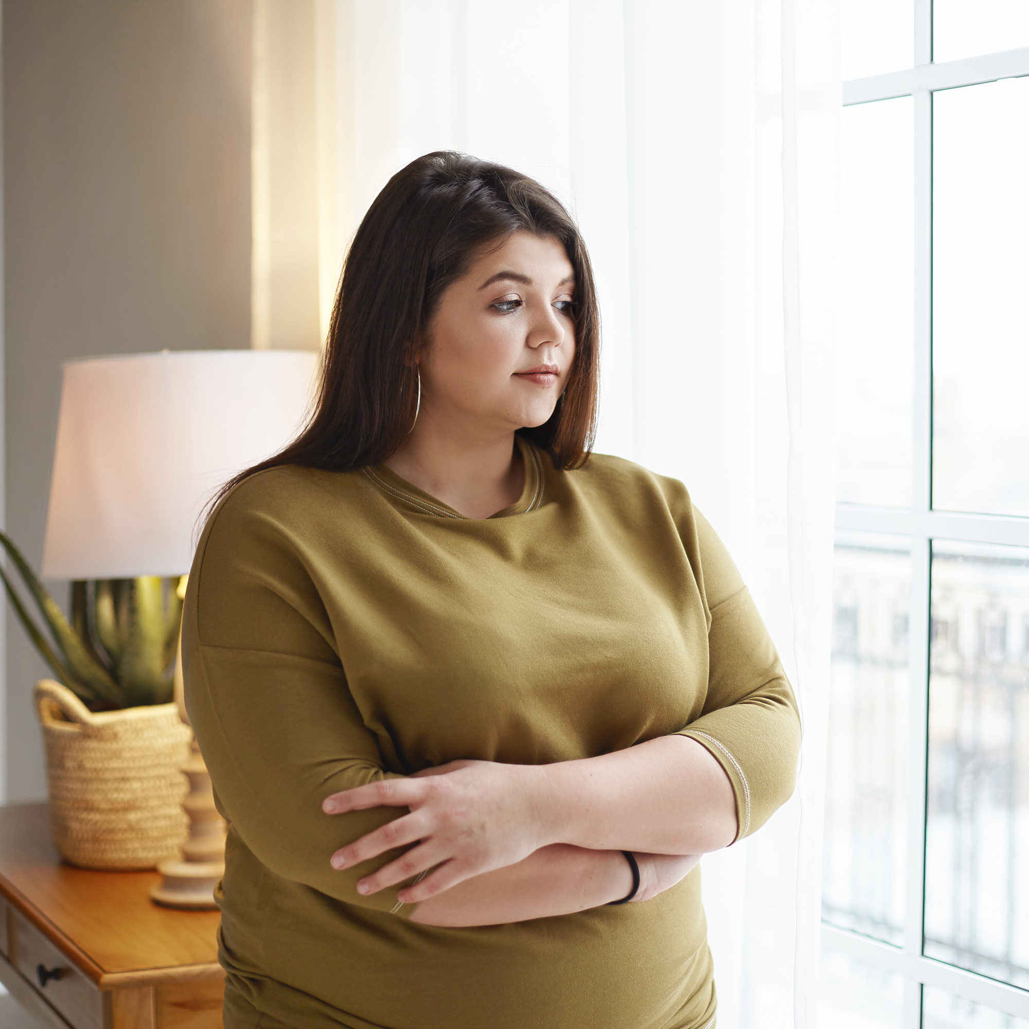 一名体重超标的女性站在家里的窗前，双臂交叉放在胸前