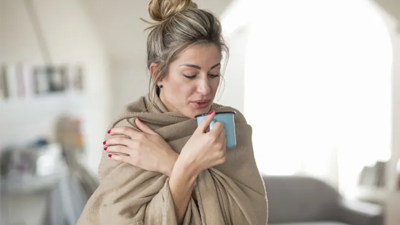 感觉冷的女人用毯子裹着，喝着茶。