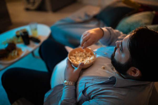 超重的人坐在沙发上看电视和吃爆米花