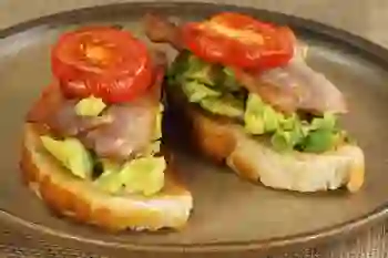 Closeup of two pieces of avocado toast topped with bacon and tomato sit on a plate.