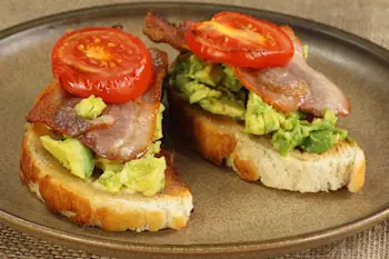 Closeup of two pieces of avocado toast topped with bacon and tomato sit on a plate.