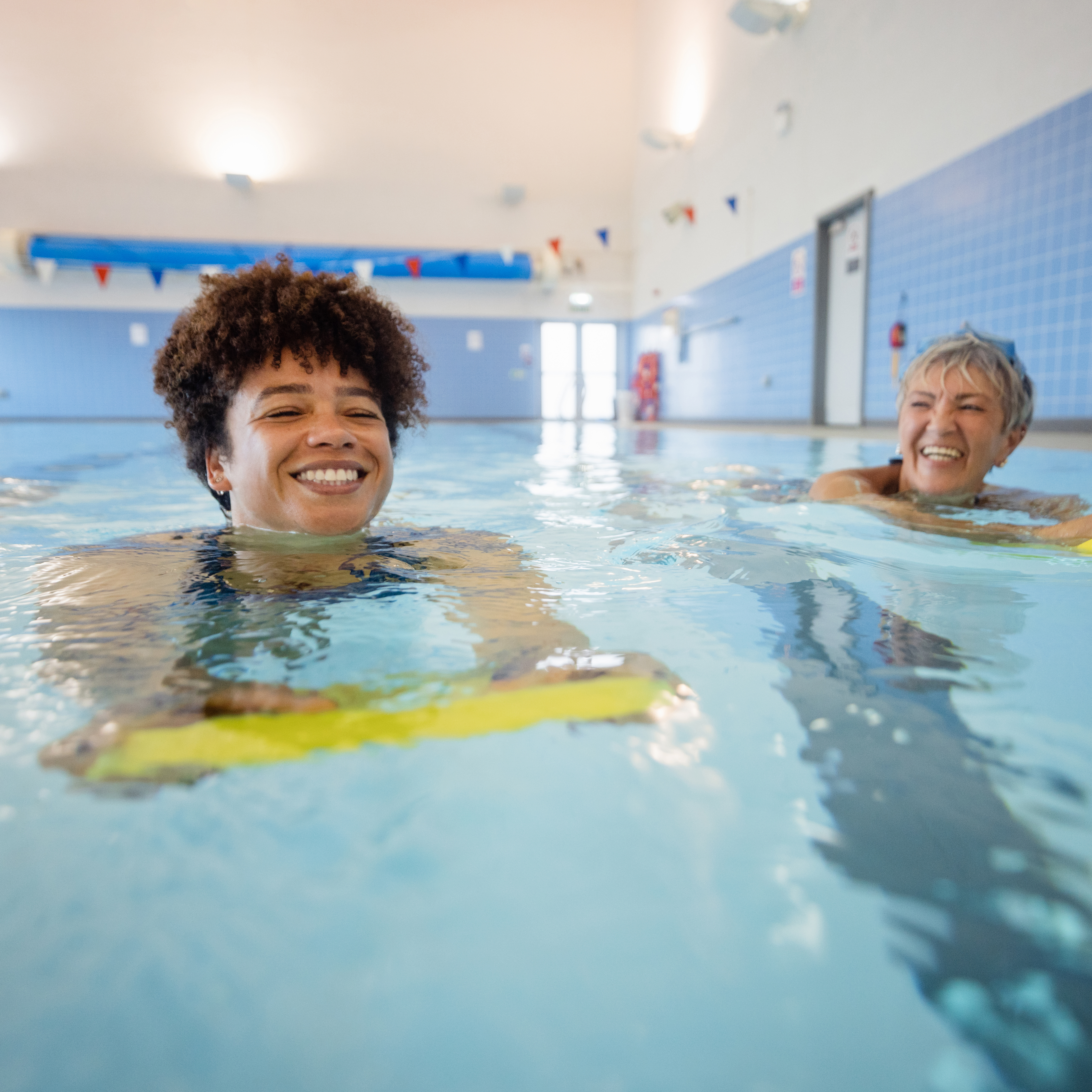 两名妇女在室内游泳池中用踢腿板游泳时微笑着。