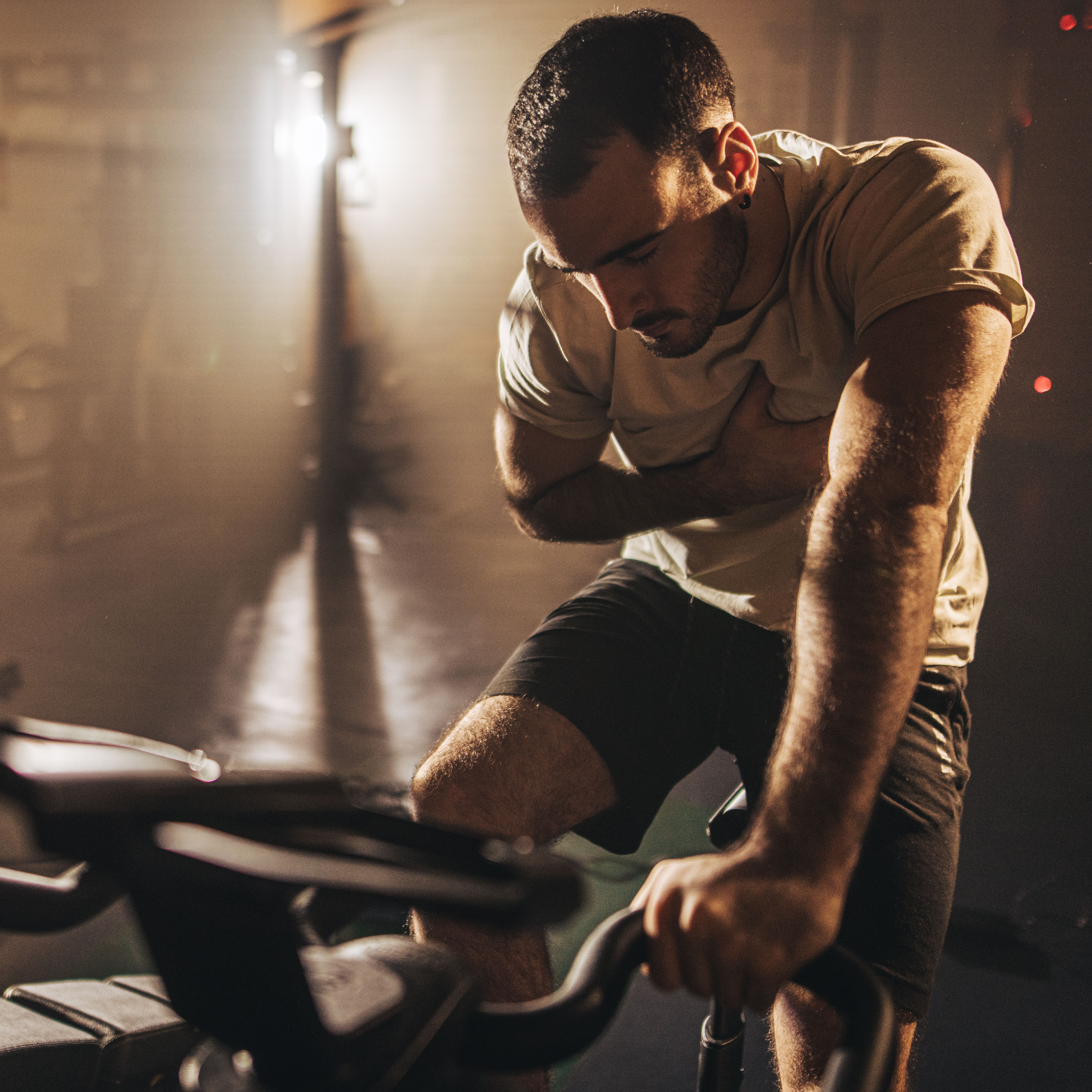 专注的健身男子独自在健身房的固定健身自行车上做心血管锻炼。他把手放在心脏上，看起来很痛苦。