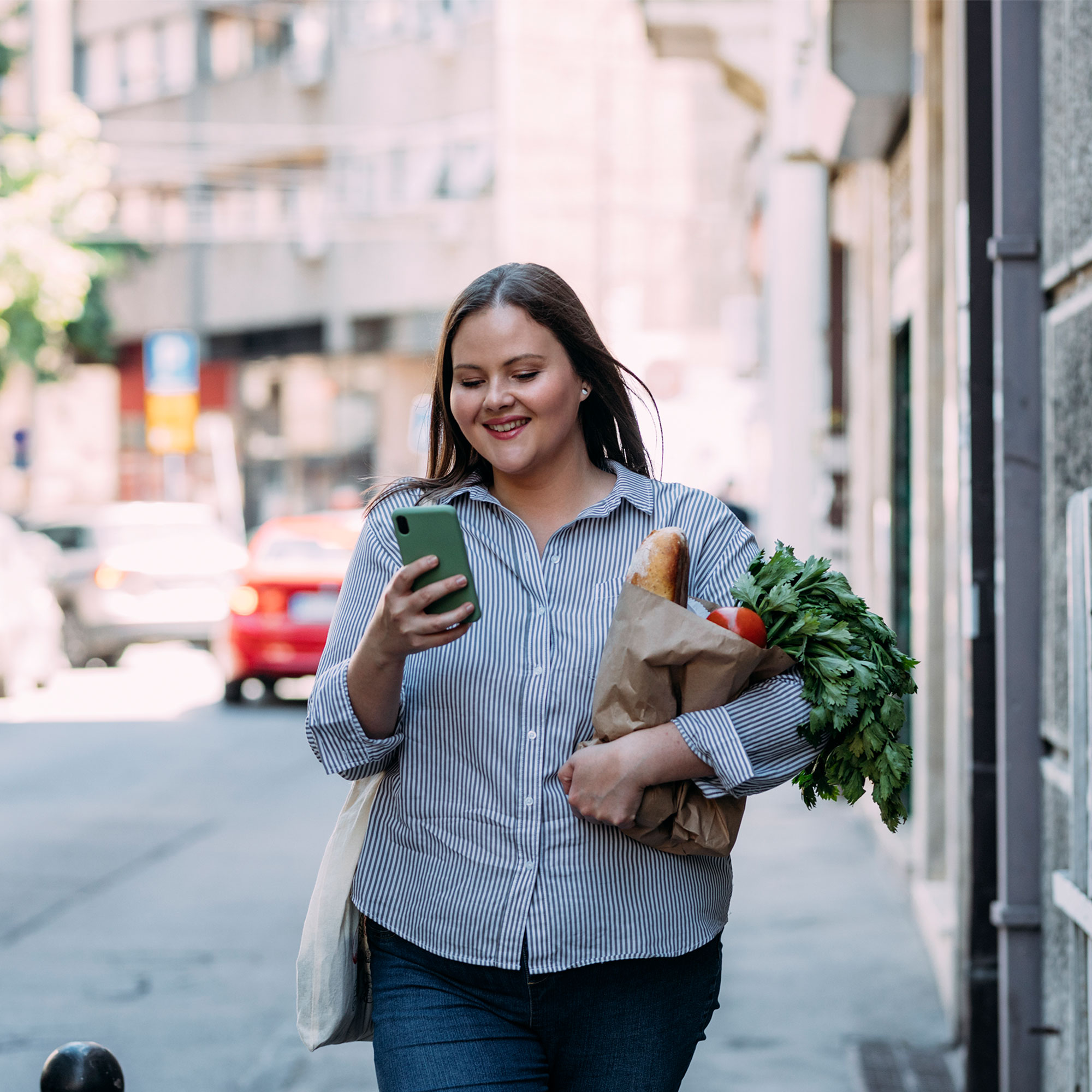 一个女人拿着杂货走在路上，看着她的手机
