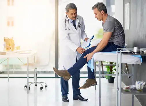 一名男子坐在医生办公室的担架上向医生解释他的腿疼。