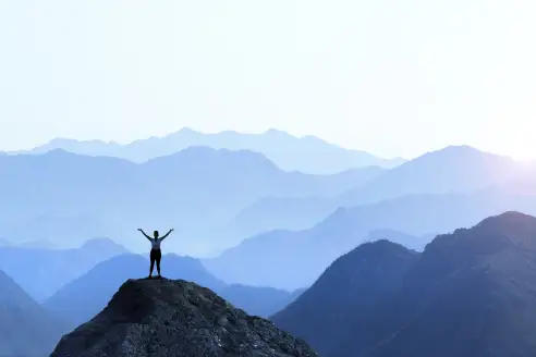 A female hiker stands on top of a rocky promontory and holds up her arms in celebration in front of a series of mountain ridges that seem to disappear into the distance as the haze obscures the details creating a graphic outline of the various layers of ri