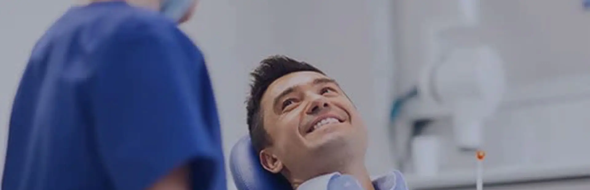 Un hombre de unos 30 años sentado en la silla del dentista sonríe durante una revisión mientras su dentista le explica que el adhesivo para prótesis dentales de Kukident ayuda a reducir las llagas.