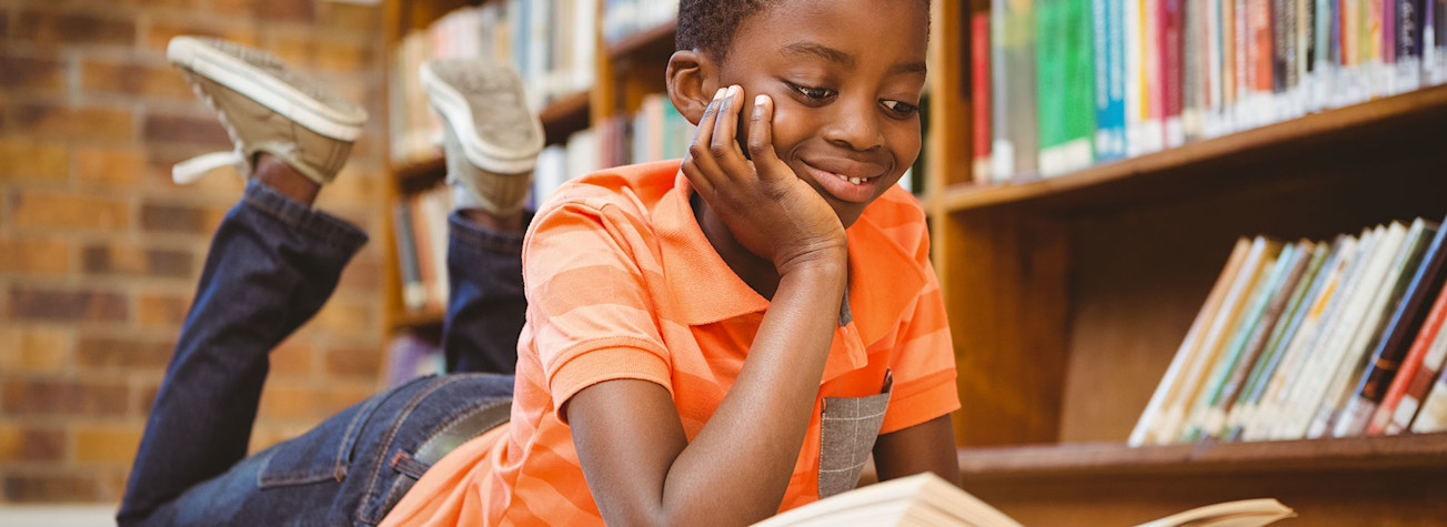 Boy lying on the floor in a library, reading a book.