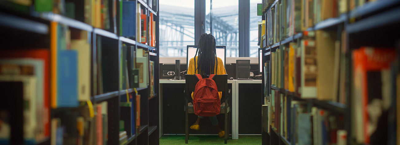 Student zitten achter een computer, gefotografeerd op de rug, door een lange gang gevormd door boekenkasten in een bibliotheek. 