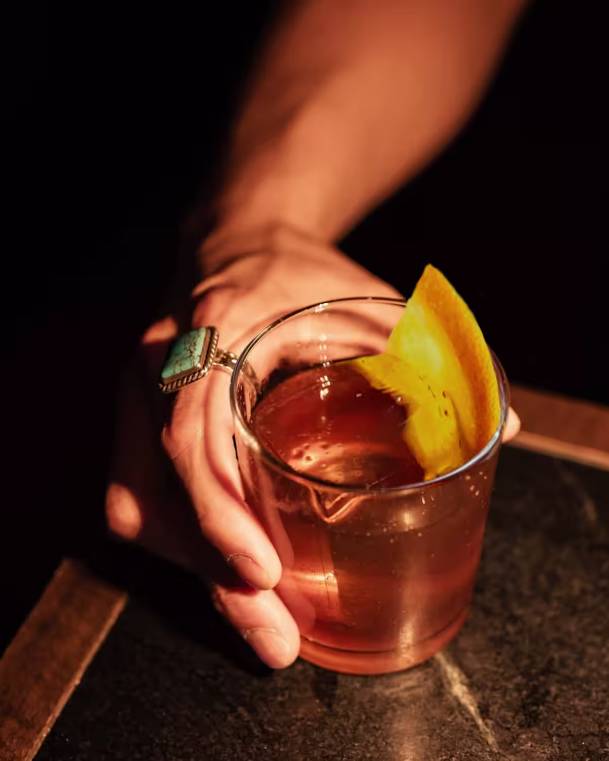 A hand wearing a turquoise ring on the pointer finger holding a rocks glass of a Coffee Negroni cocktail with orange garnish.