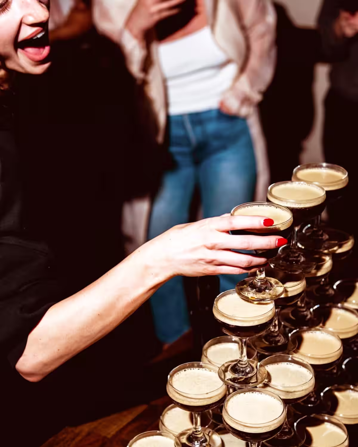 A woman with a big smile grabbing an Espresso Mint-tini cocktail from a stacked martini glass tower in a party setting.