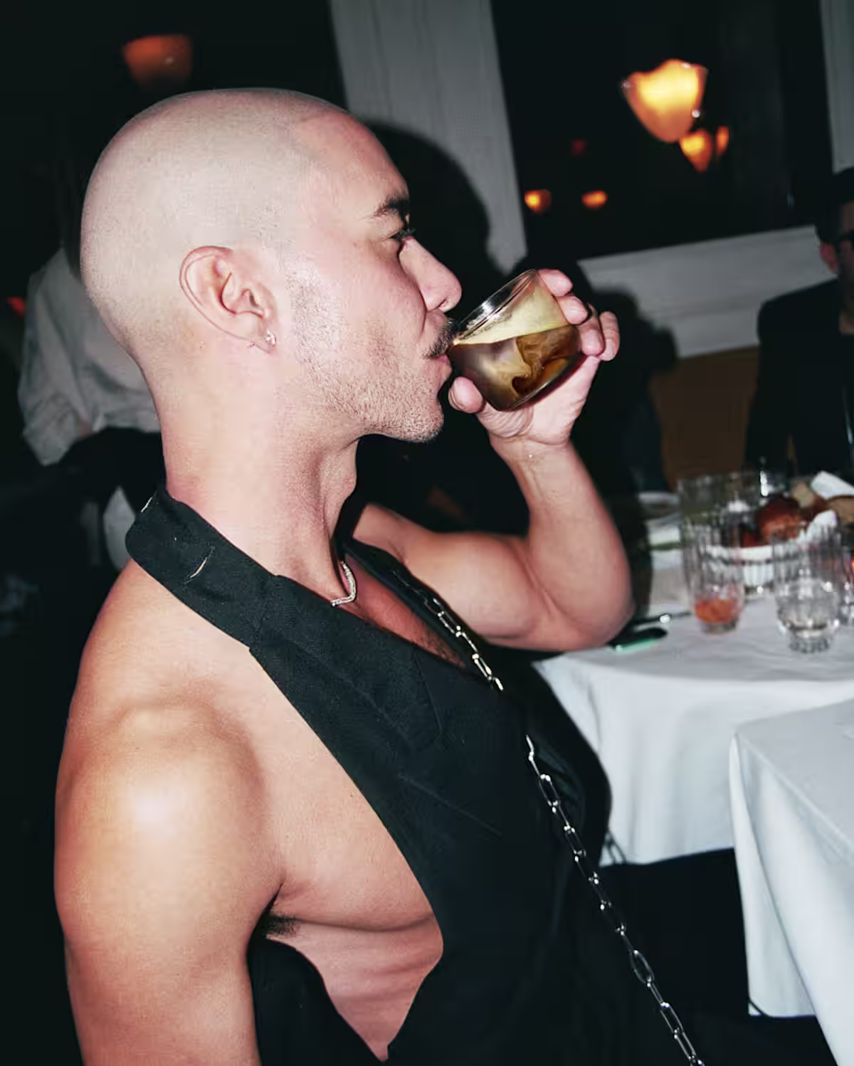 A man with a shaved head wearing a tuxedo vest drinking a White Russian cocktail in a restaurant