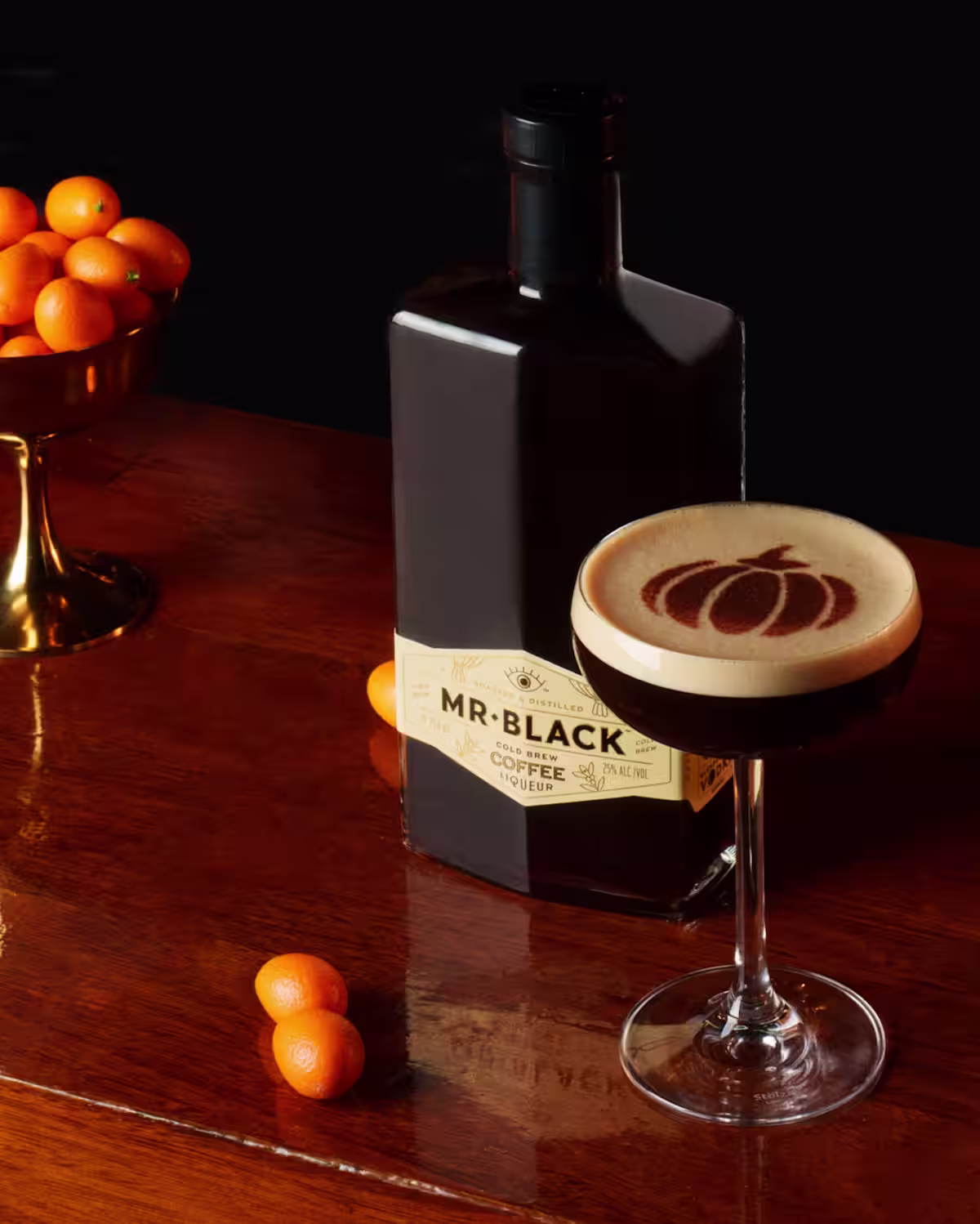 A Pumpkin Spice Espresso Martini with a pumpkin latte art decor on top next to a bottle of Mr Black Coffee Liqueur and a bowl of orange kumquats.