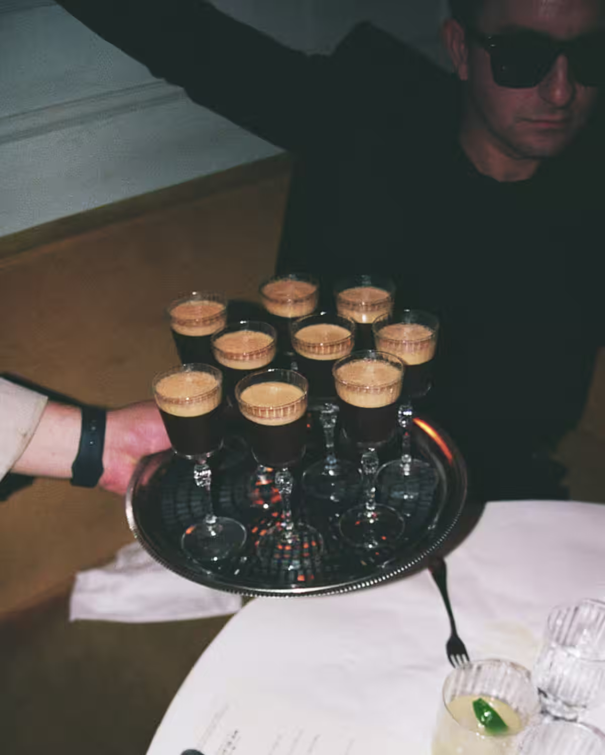 A tray of Espresso Martini cocktails being handed to a man wearing all black with dark sunglasses in a dining room setting.