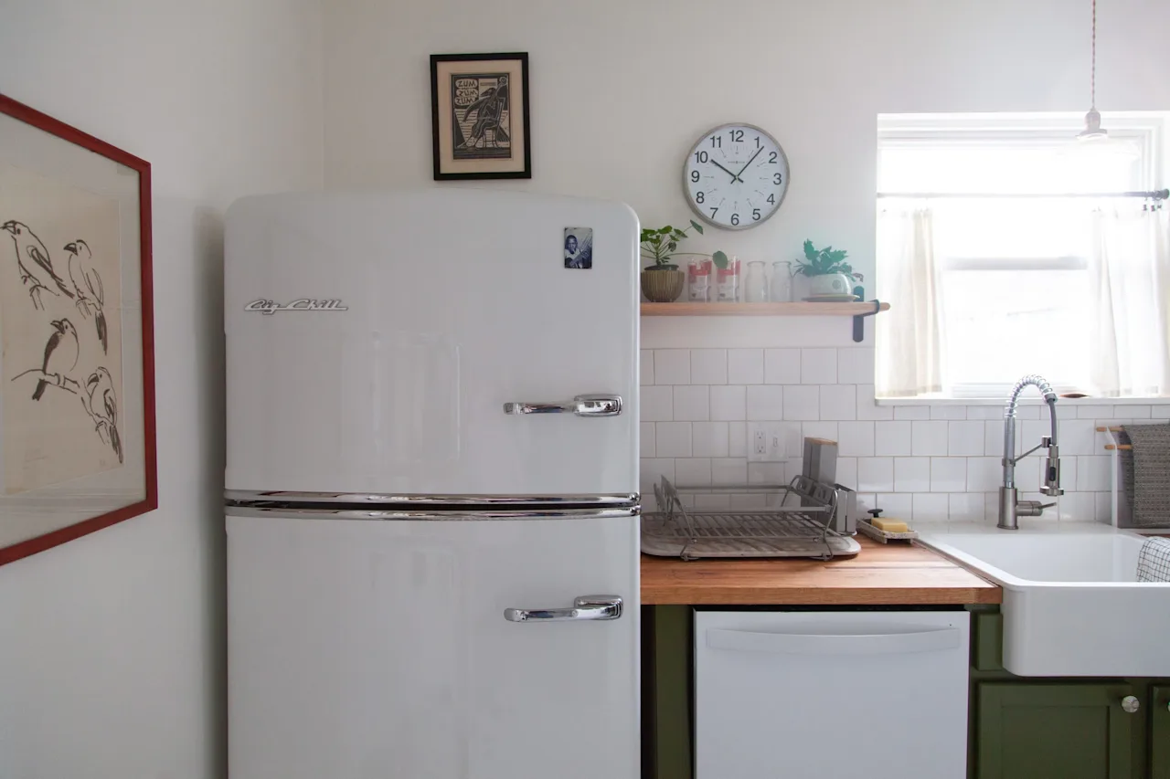 Compact kitchen with a white retro refrigerator, white subway tile, an open wood shelf, and a farmhouse sink with butcher-block counter.