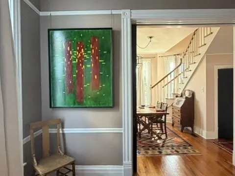 Sitting area with an abstract green painting and wainscoting, looking through to a dining room and staircase.