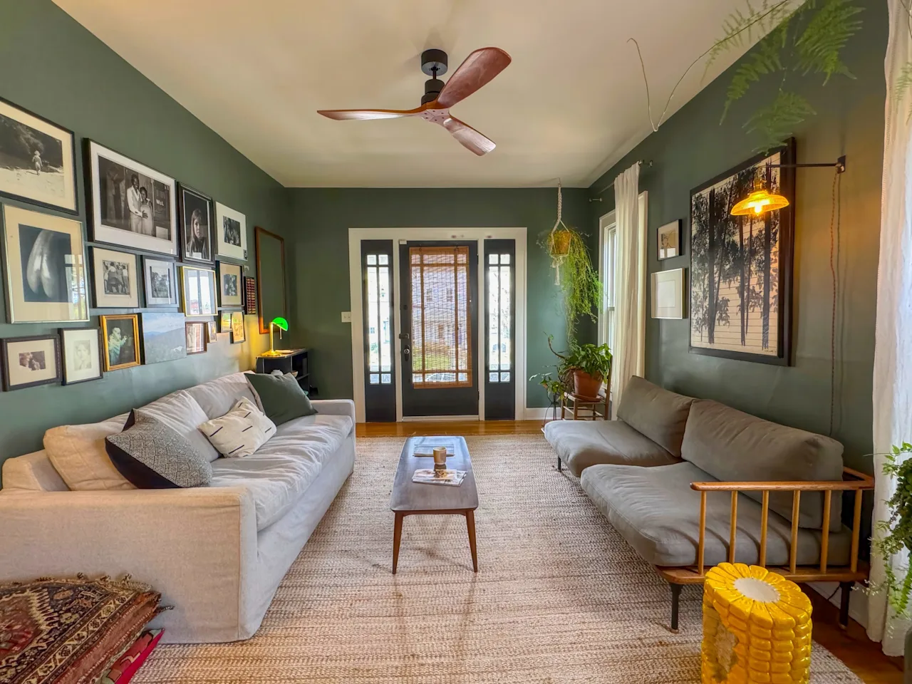 Green-walled living room with a ceiling fan, a gallery wall of framed art, two sofas, and a mid-century coffee table on a jute rug.