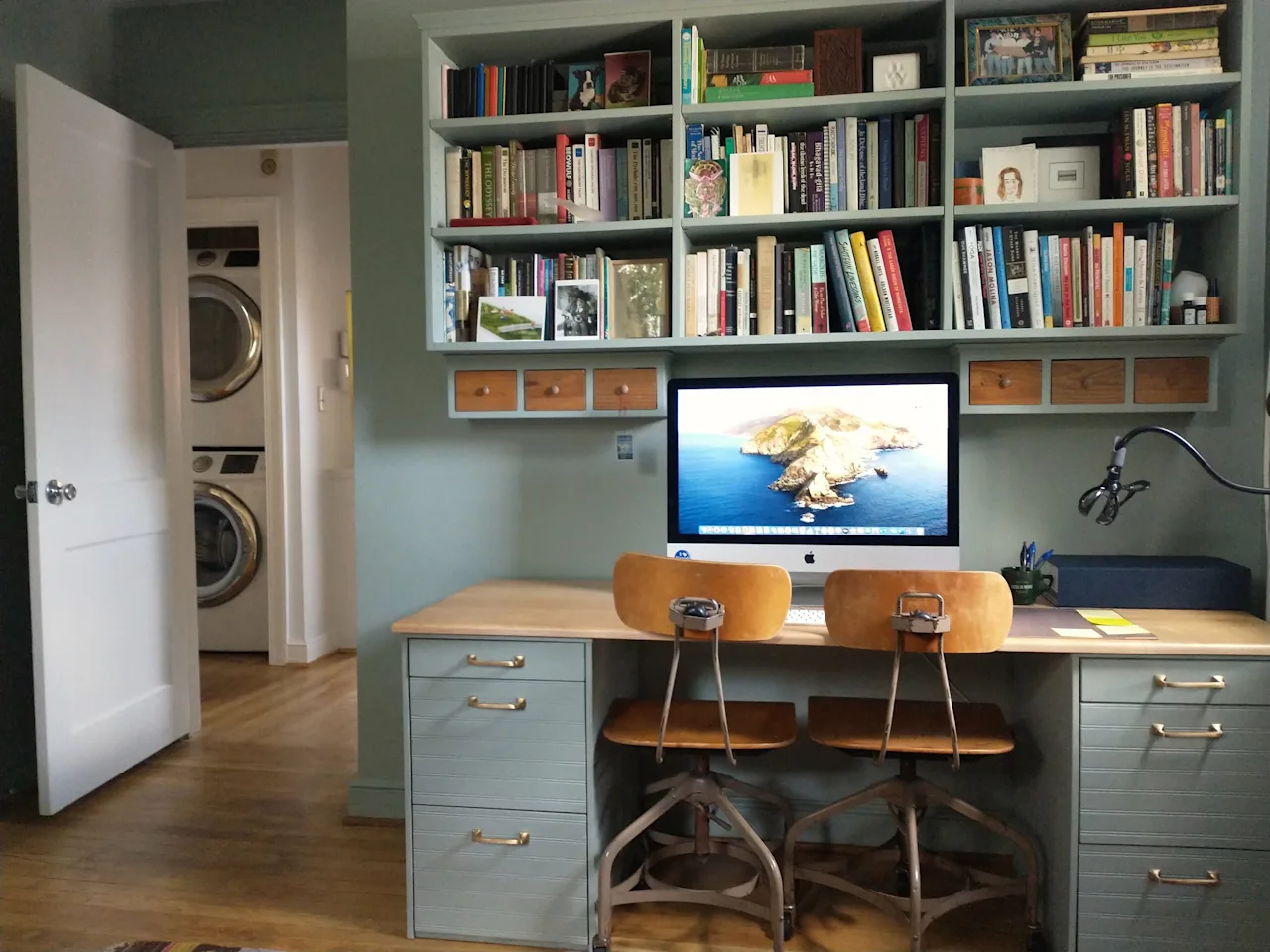 Home office with a built-in wood desk and stools beside a laundry nook with a stacked washer and dryer.